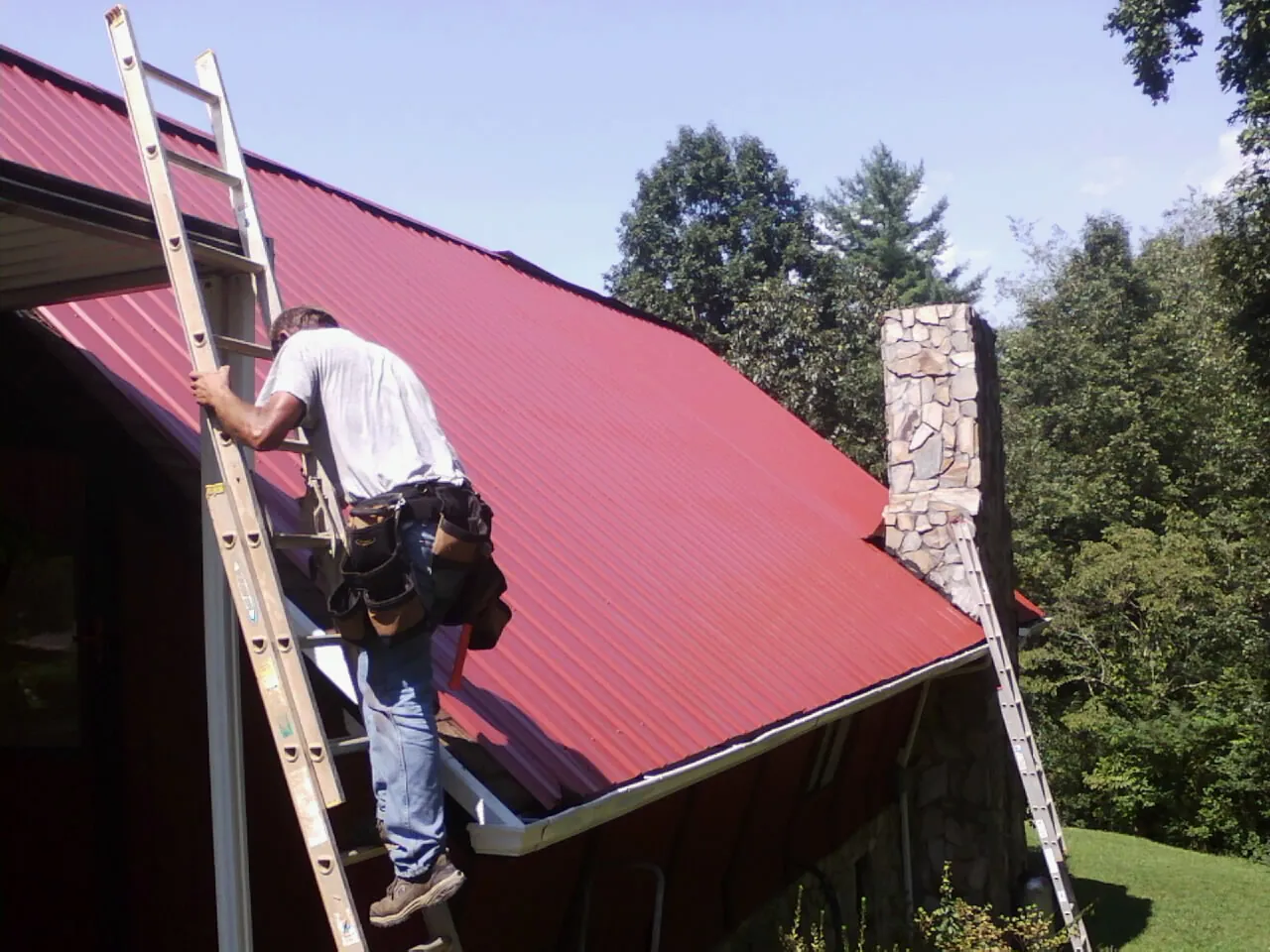 Roofer on a ladder beside a red metal roof and stone chimney — Soffit & Fascia Repair by Roofing Brothers in Bryantown